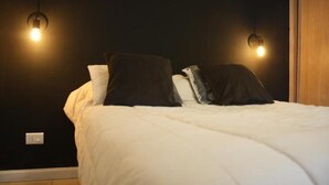 A bed with white bedding and two black pillows, two wall-mounted light fixtures, and a wooden door. - Charming 1-bedroom apartment with WiFi, AC in fabulous GFJ (GFJ)