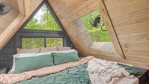 A cozy bedroom with a sloped ceiling, a large window, and a bed with green and beige bedding. - Luxury A-Frame: Pets, Pickleball & Peloton w/Views (Mineral Bluff)