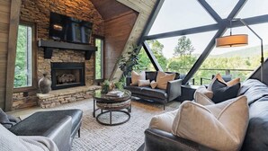 A cozy living room with a stone fireplace, leather sofas, and a large window offering a scenic view. - Luxury A-Frame: Pets, Pickleball & Peloton w/Views (Mineral Bluff)