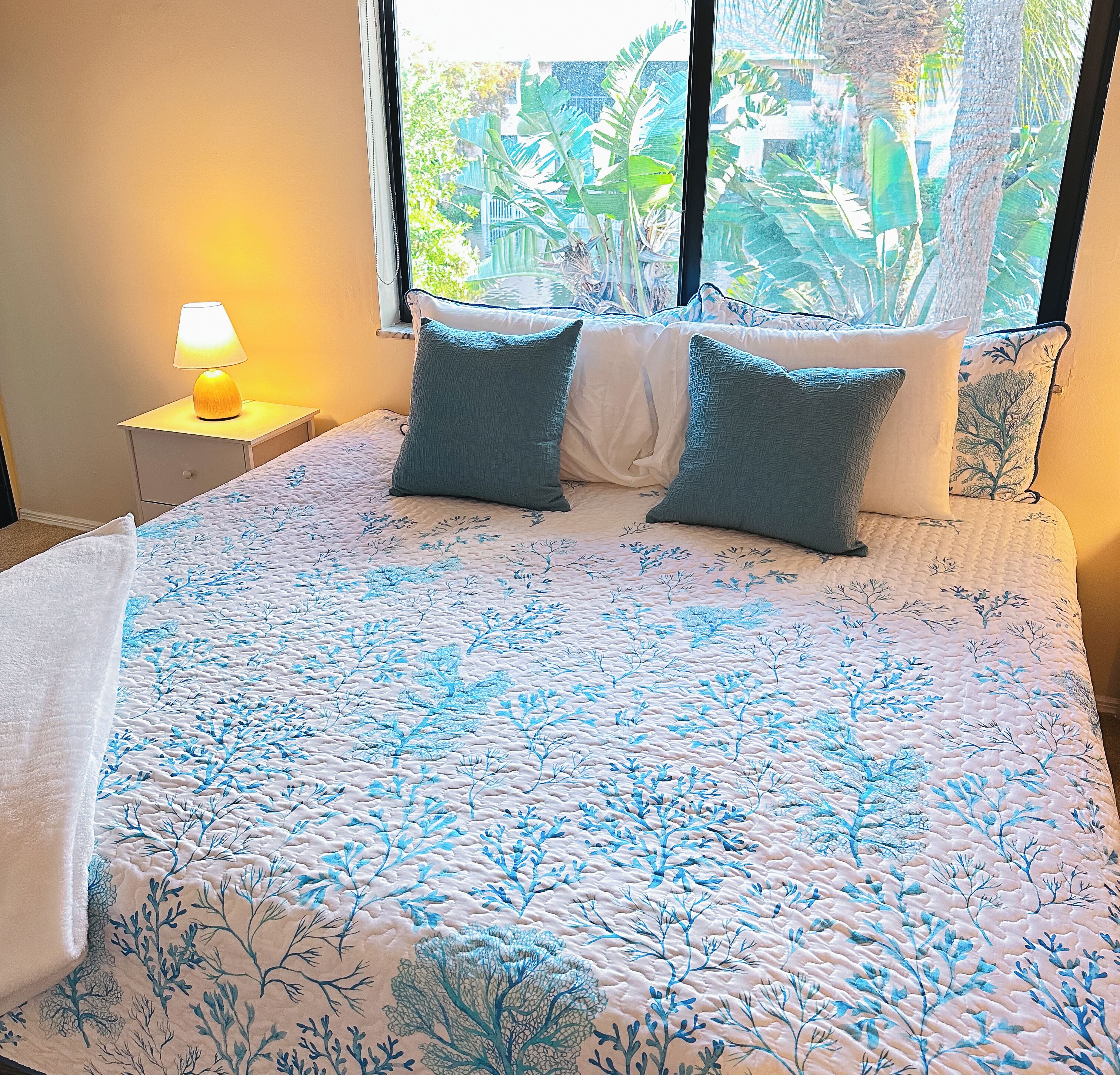 Una cama con un edredón azul con motivos florales, dos almohadas, una mesita de noche con una lámpara y una ventana con vistas a zonas verdes.