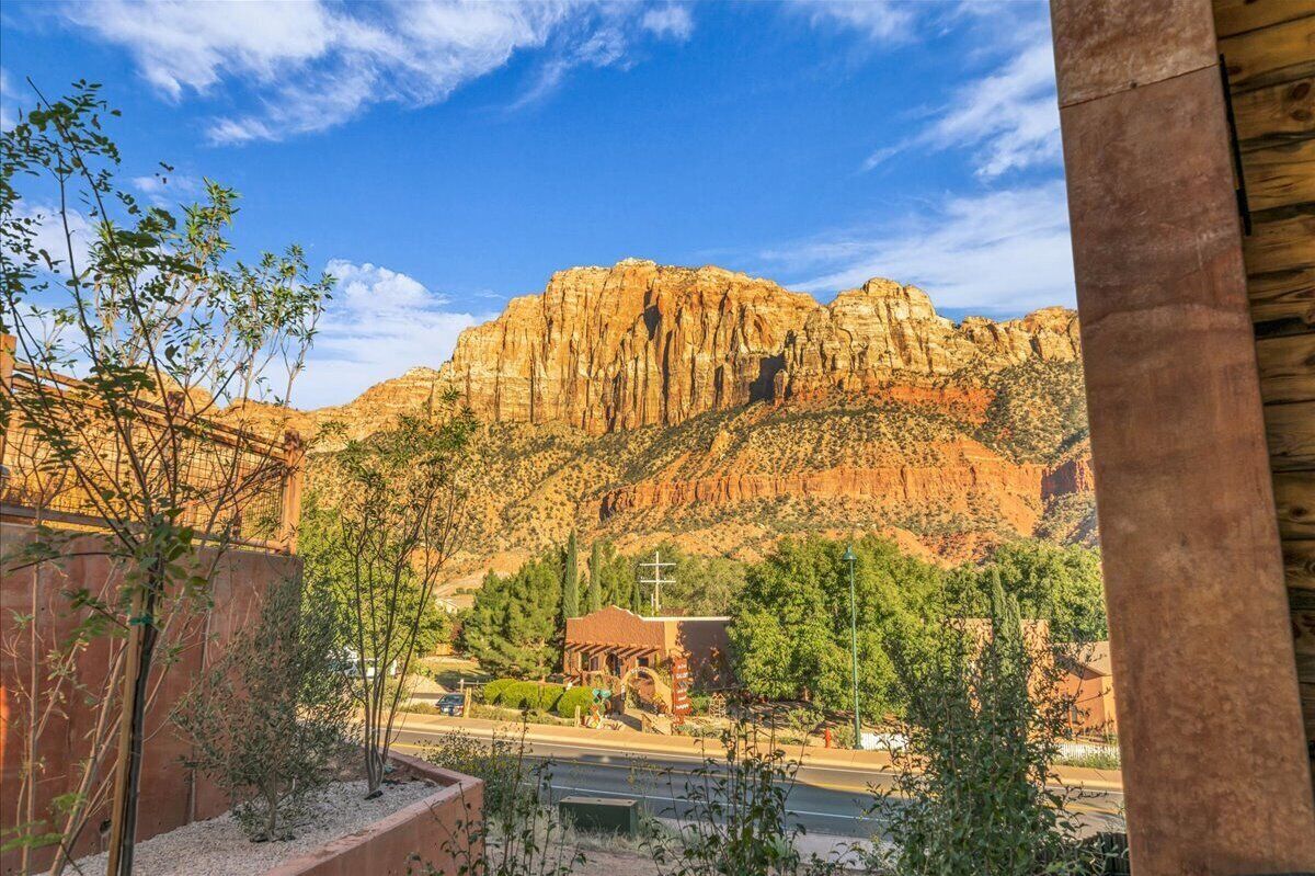 Vista de un paisaje montañoso con un pequeño edificio en primer plano, posiblemente una capilla o lugar religioso, rodeado de vegetación y un cielo despejado.