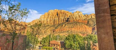 Vista de un paisaje montañoso con un pequeño edificio en primer plano, posiblemente una capilla o lugar religioso, rodeado de vegetación y un cielo despejado.