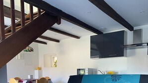 Una sala de estar con una mesa de centro de vidrio, un sofá azul, sillas de madera, una escalera y una zona de cocina con armarios y un microondas.