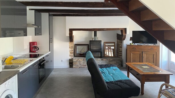 Una cocina moderna con lavavajillas, una cafetera roja y lavadora. Hay una zona de estar acogedora con un sofá negro y una mesa de madera. Un televisor está montado en la pared, y se ve una chimenea de piedra al fondo.