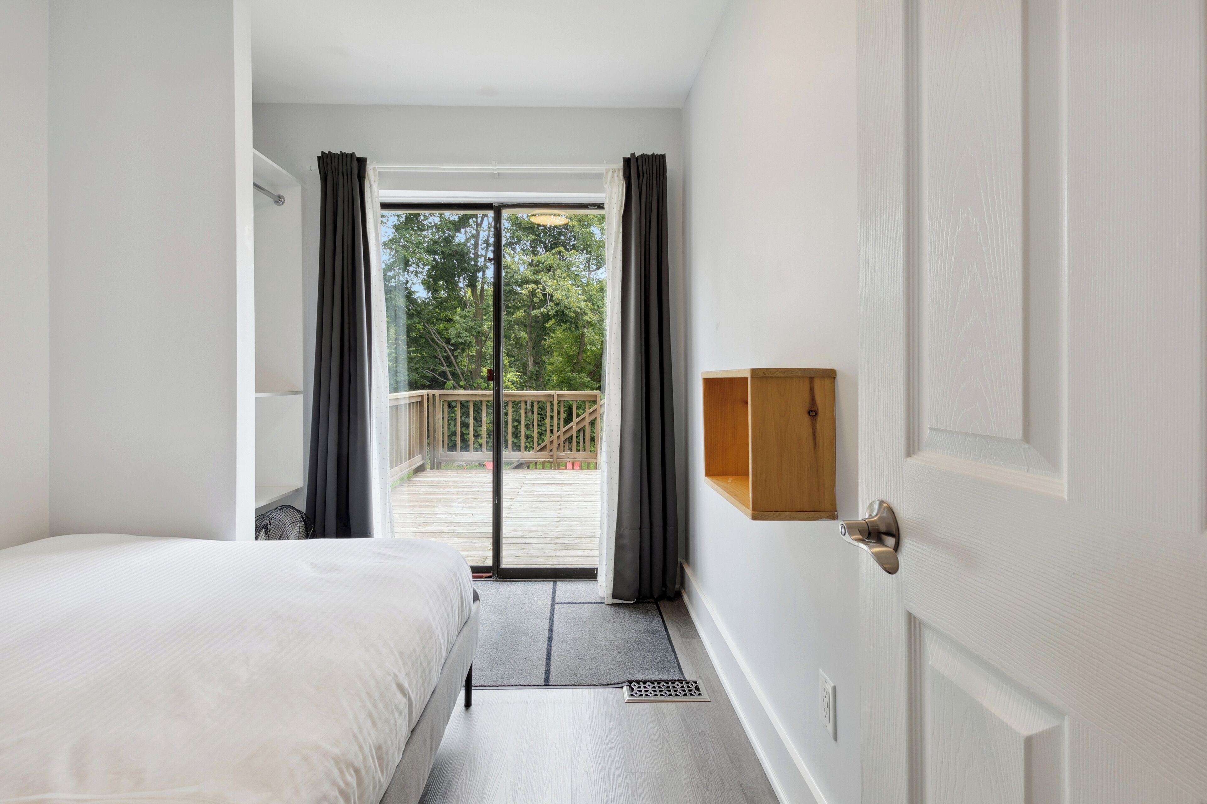 A bedroom with a bed, a wooden shelf, a door, and a view of a balcony with a railing.