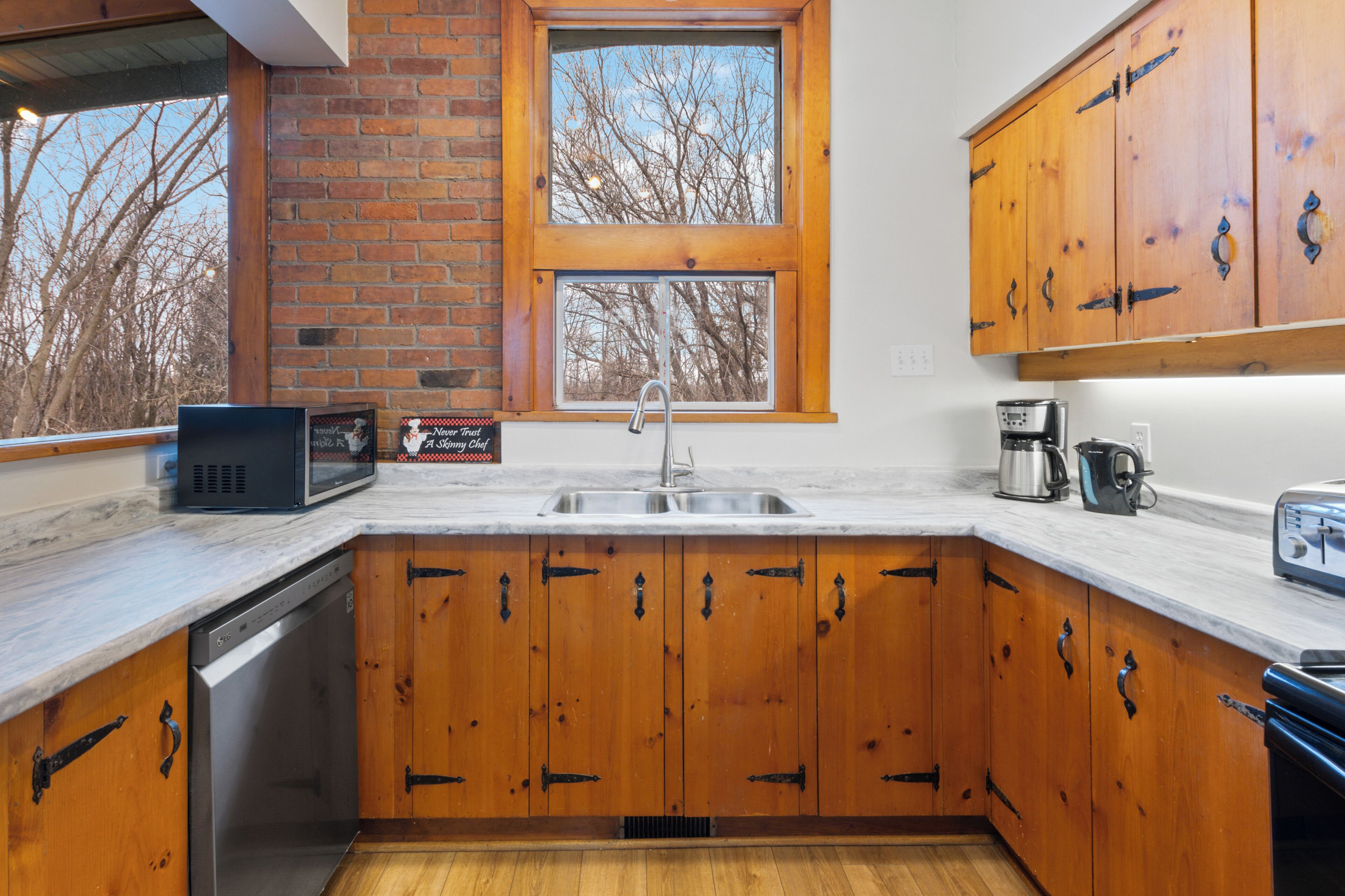 A kitchen with wooden cabinets, a dishwasher, a sink, and a window with a view of trees.