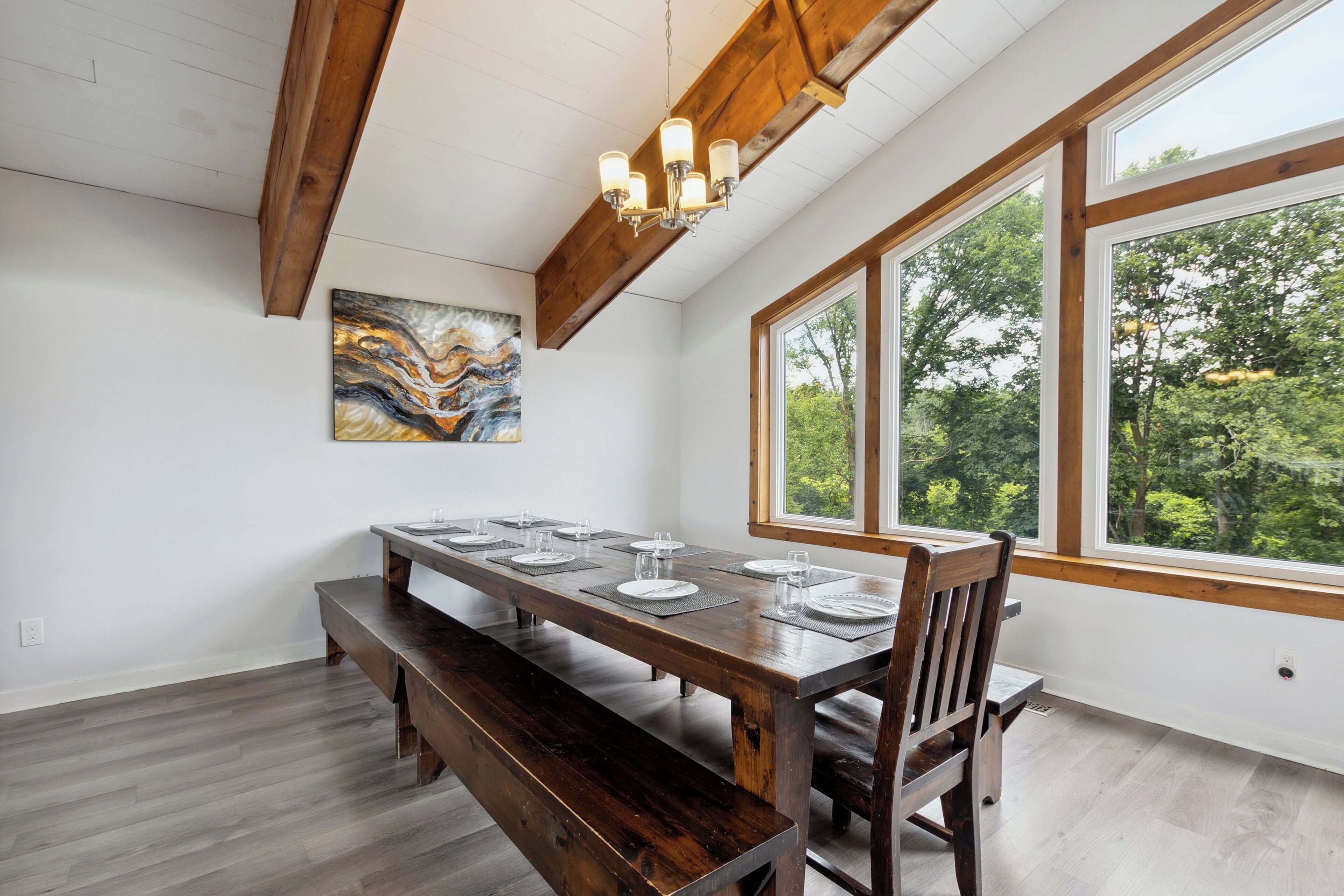 Un coin repas avec une table en bois dressée pour six personnes, un banc et une grande fenêtre donnant sur les arbres.