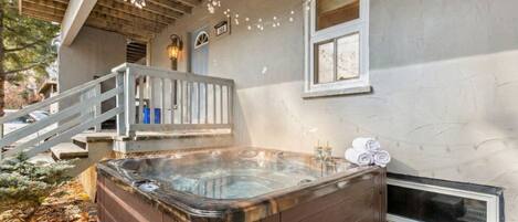 A hot tub outside a house with a wooden deck and stairs leading up to the entrance.