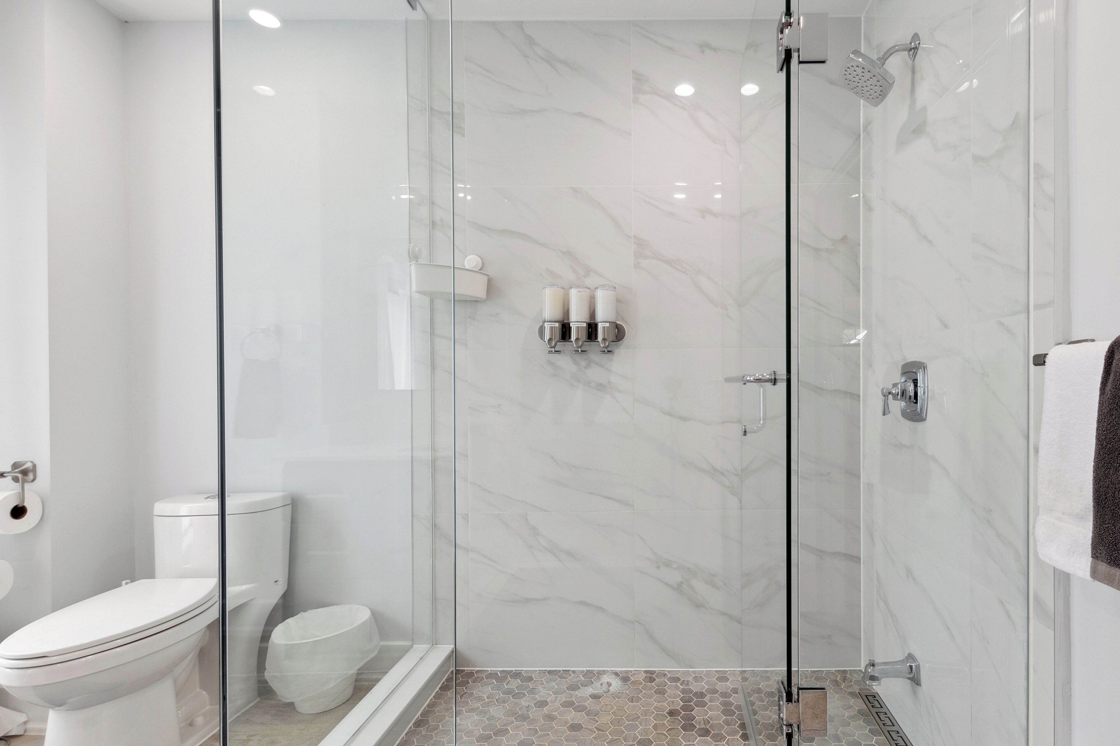 A modern bathroom with a glass-enclosed shower, a toilet, and a sink with three bottles of soap.