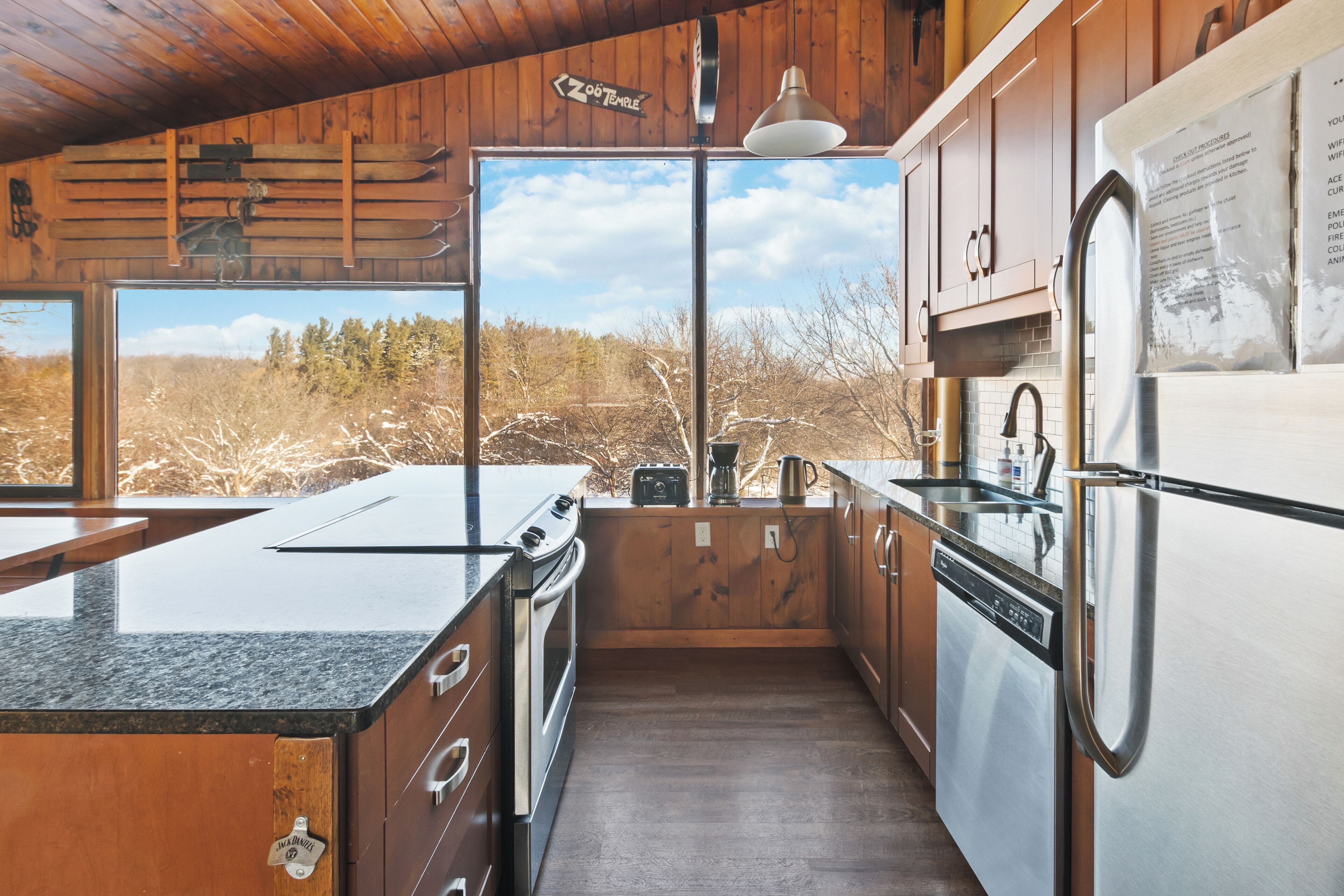 A compact kitchen with wooden cabinets, a built-in oven, a dishwasher, and a large window offering a view of the outdoors.