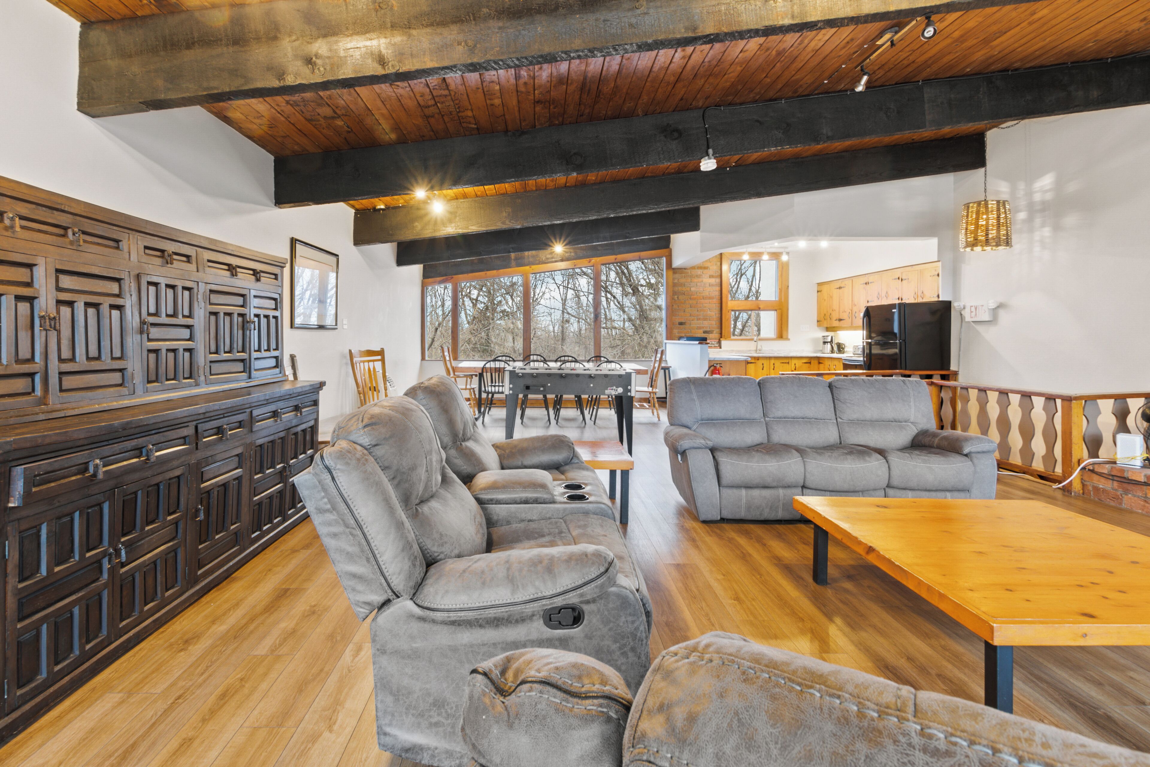 A spacious living area with a wooden ceiling, a gray sofa, a wooden dining table, and a kitchen in the background.