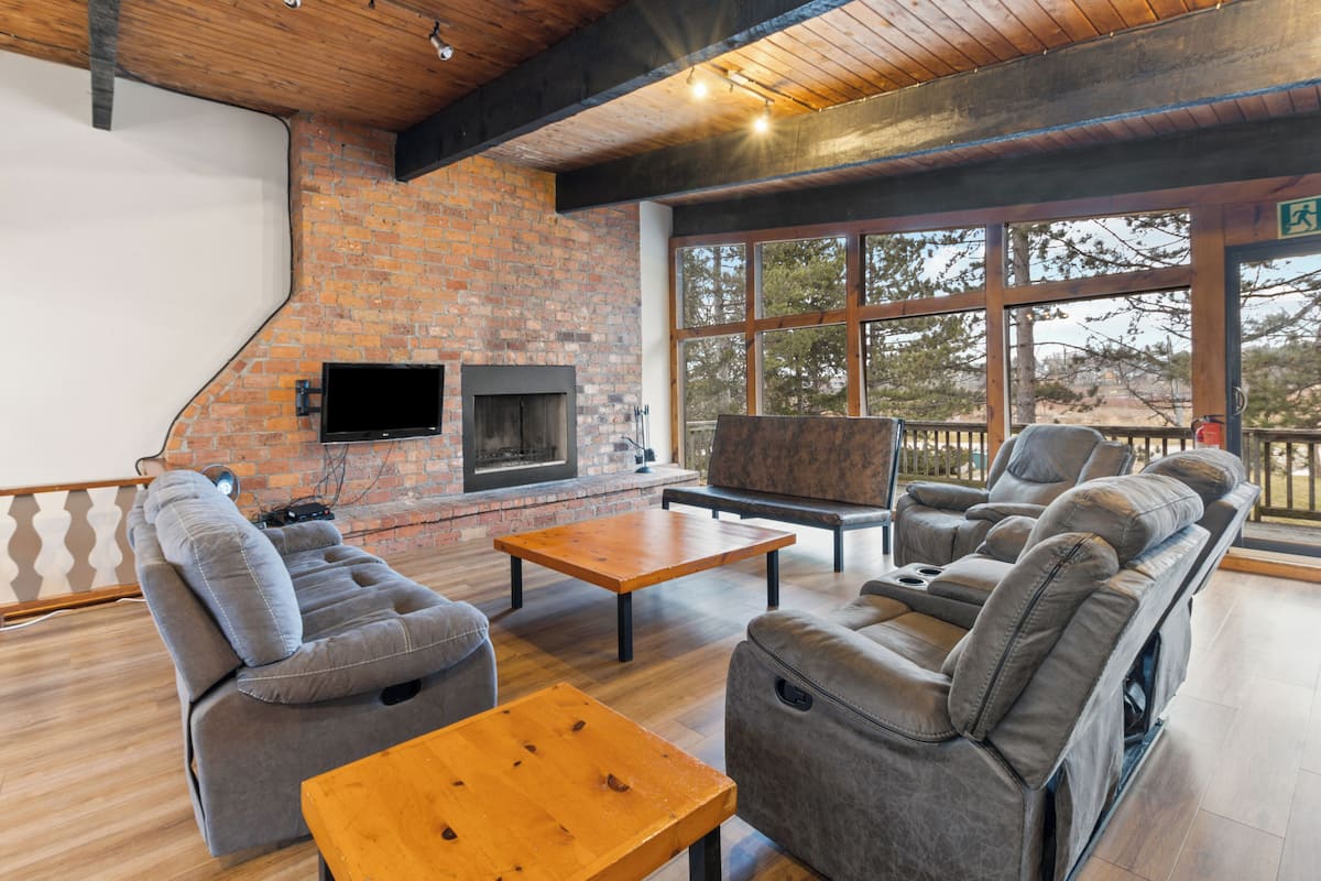 Un área de estar moderna con pared de ladrillo, chimenea, asientos de cuero y grandes ventanales que ofrecen vistas al exterior.