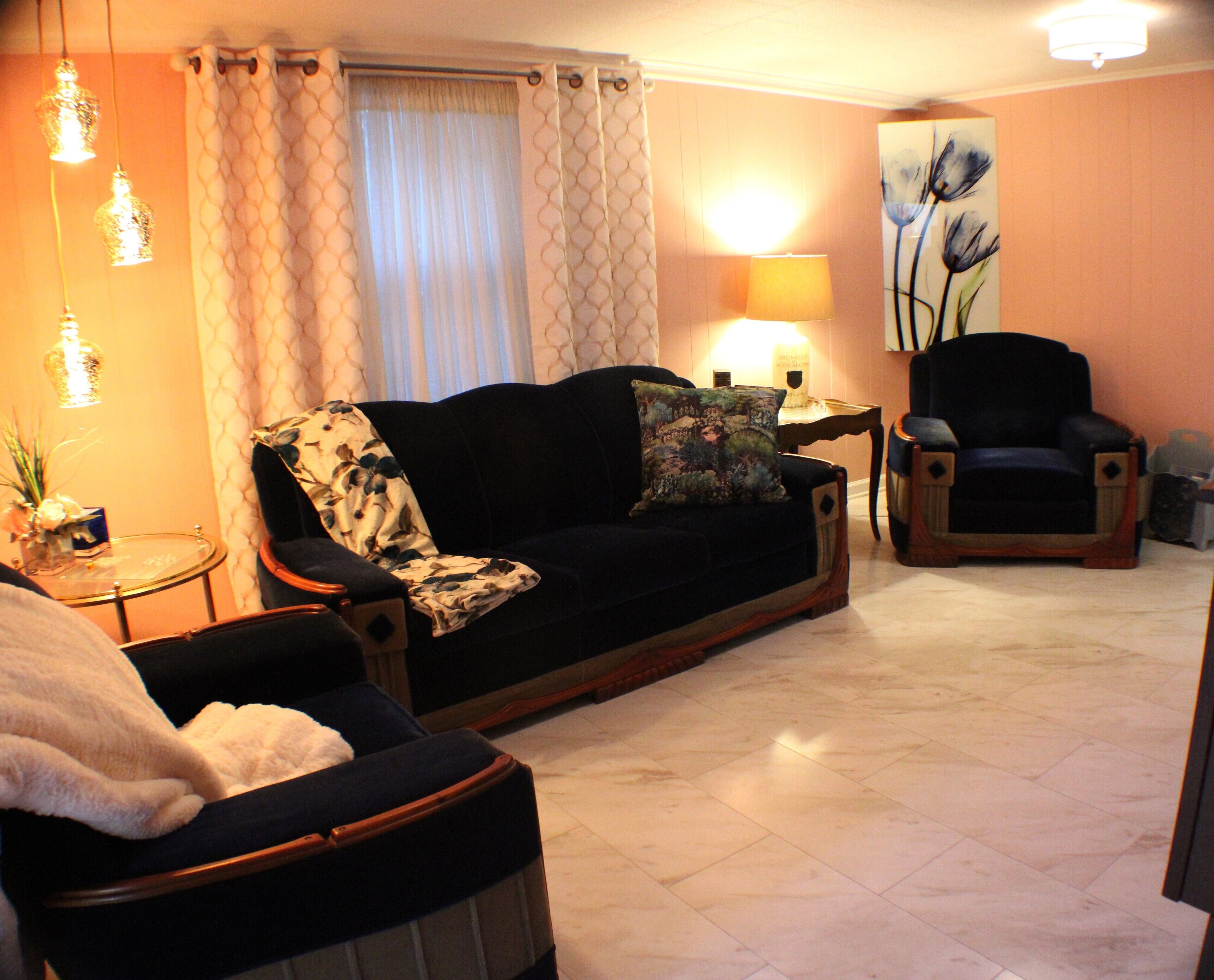 A living room with a dark sofa, a patterned throw, a side table with a lamp, a chair, and a painting on the wall.