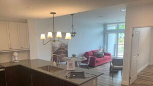 A modern kitchen with a central island, a dining area with a red sofa, and a view of the outdoors through large windows. - New The Carlyle 204 Grand Ben (Grand Bend)