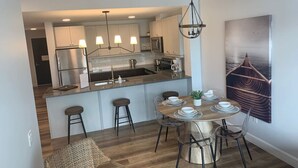 A modern kitchen with a central island, bar stools, and a dining area with a table set for four. - New The Carlyle 204 Grand Ben (Grand Bend)
