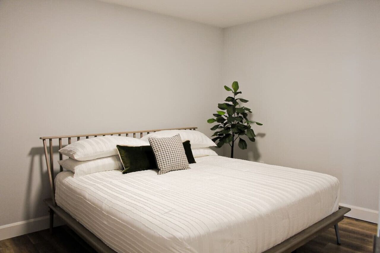 Ein ordentlich bezogenes Bett mit Holzrahmen und weißer Bettwäsche, eine Topfpflanze rechts daneben und eine schlichte weiße Wand.