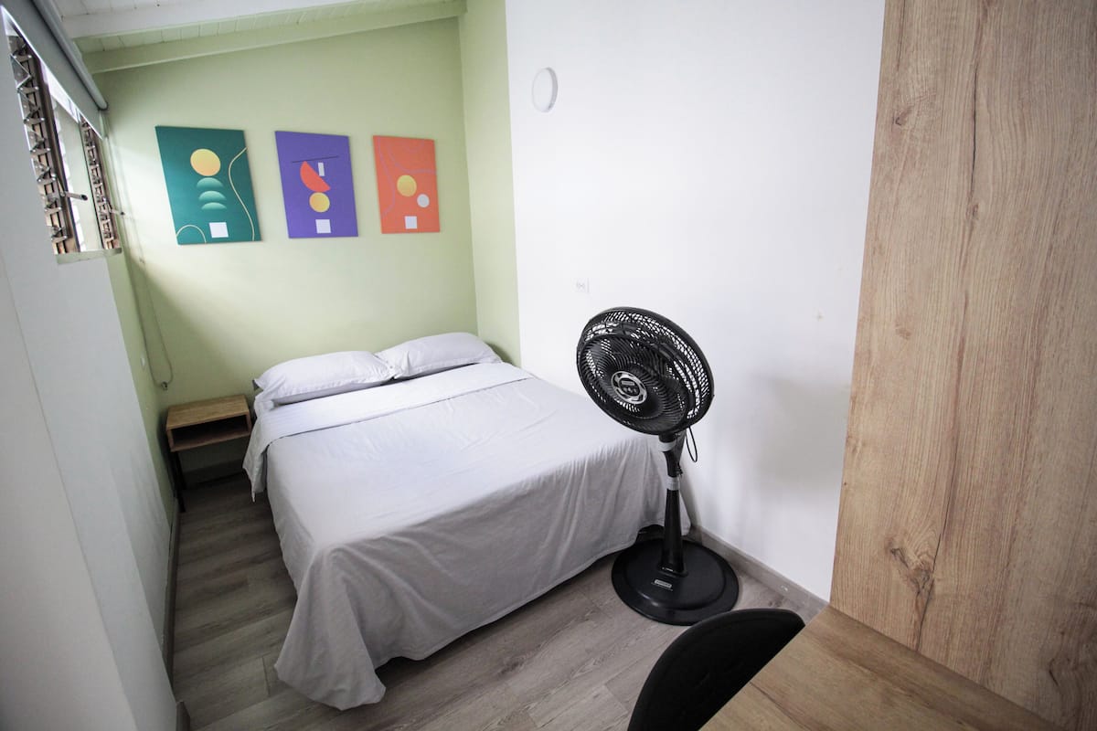 A small, narrow room with a bed, a standing fan, and three abstract paintings on the wall.