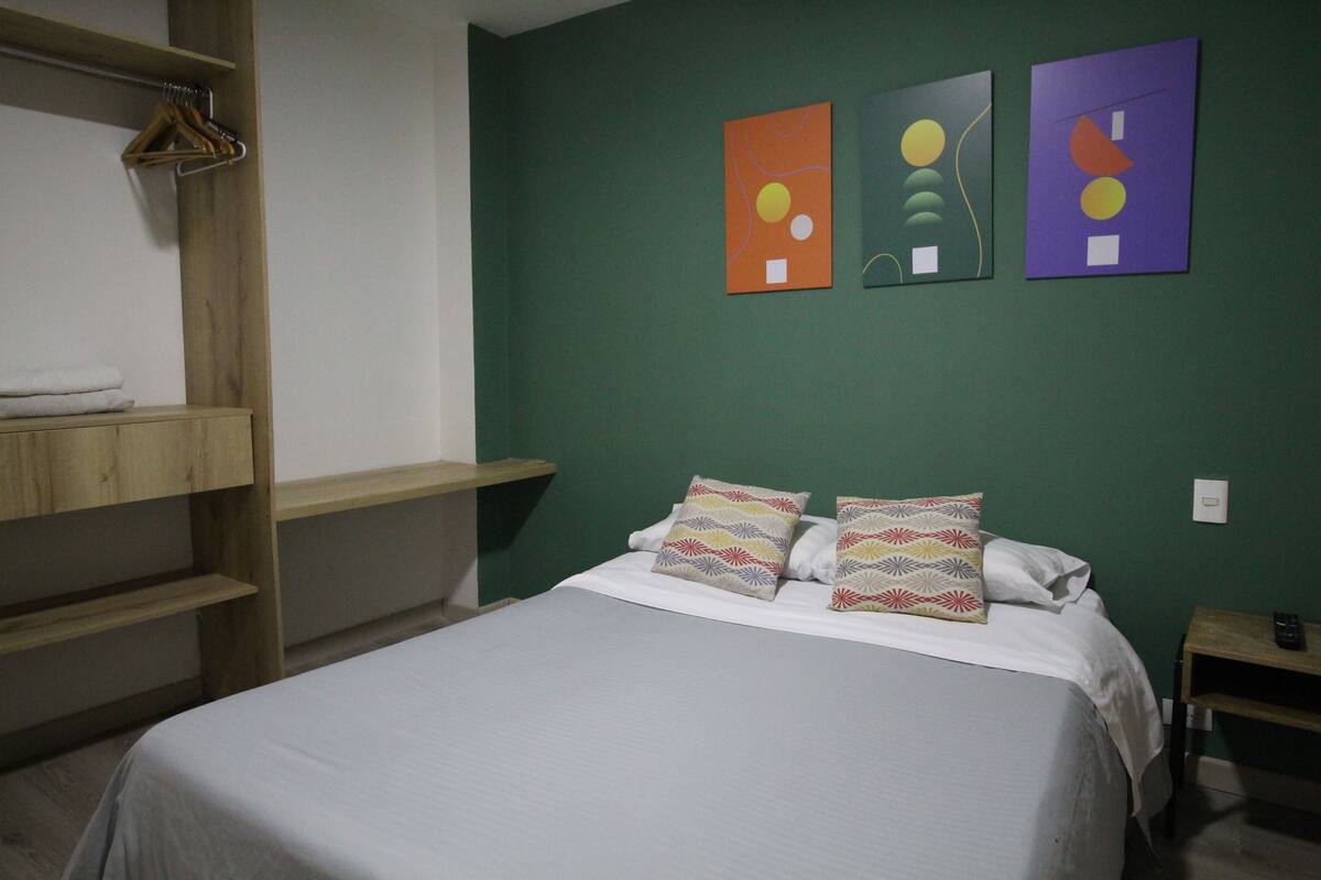 A room with a bed, a wooden shelf, and three abstract paintings on the wall.