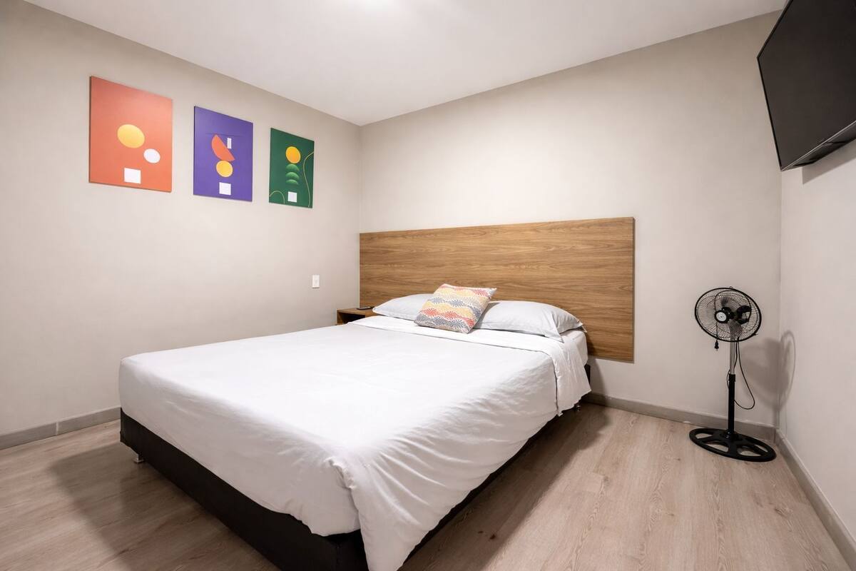 A bedroom with a bed, a nightstand, a wall-mounted TV, and a standing fan.