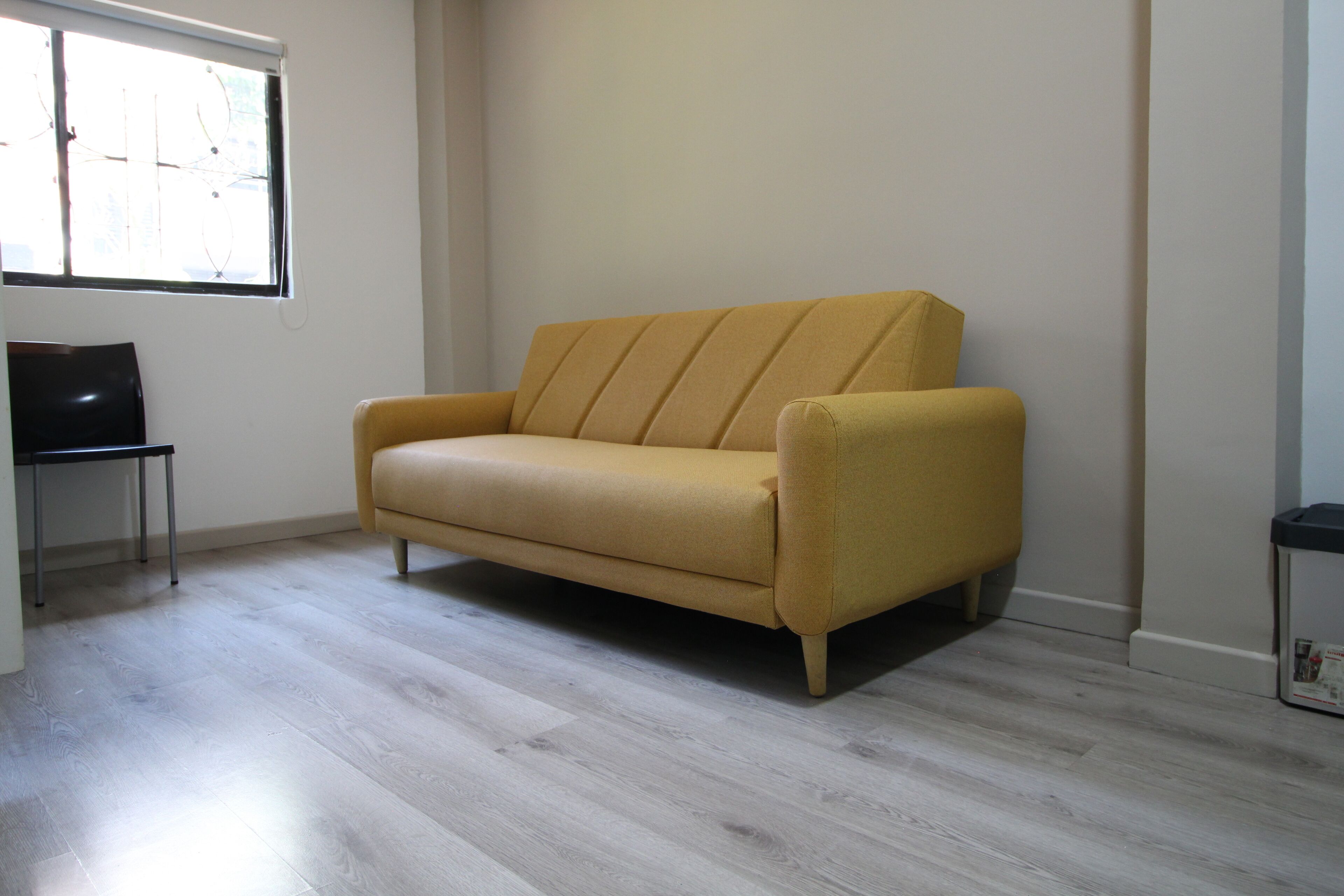 A room with a light-colored wooden floor, a yellow sofa, a small table, and a window with blinds.