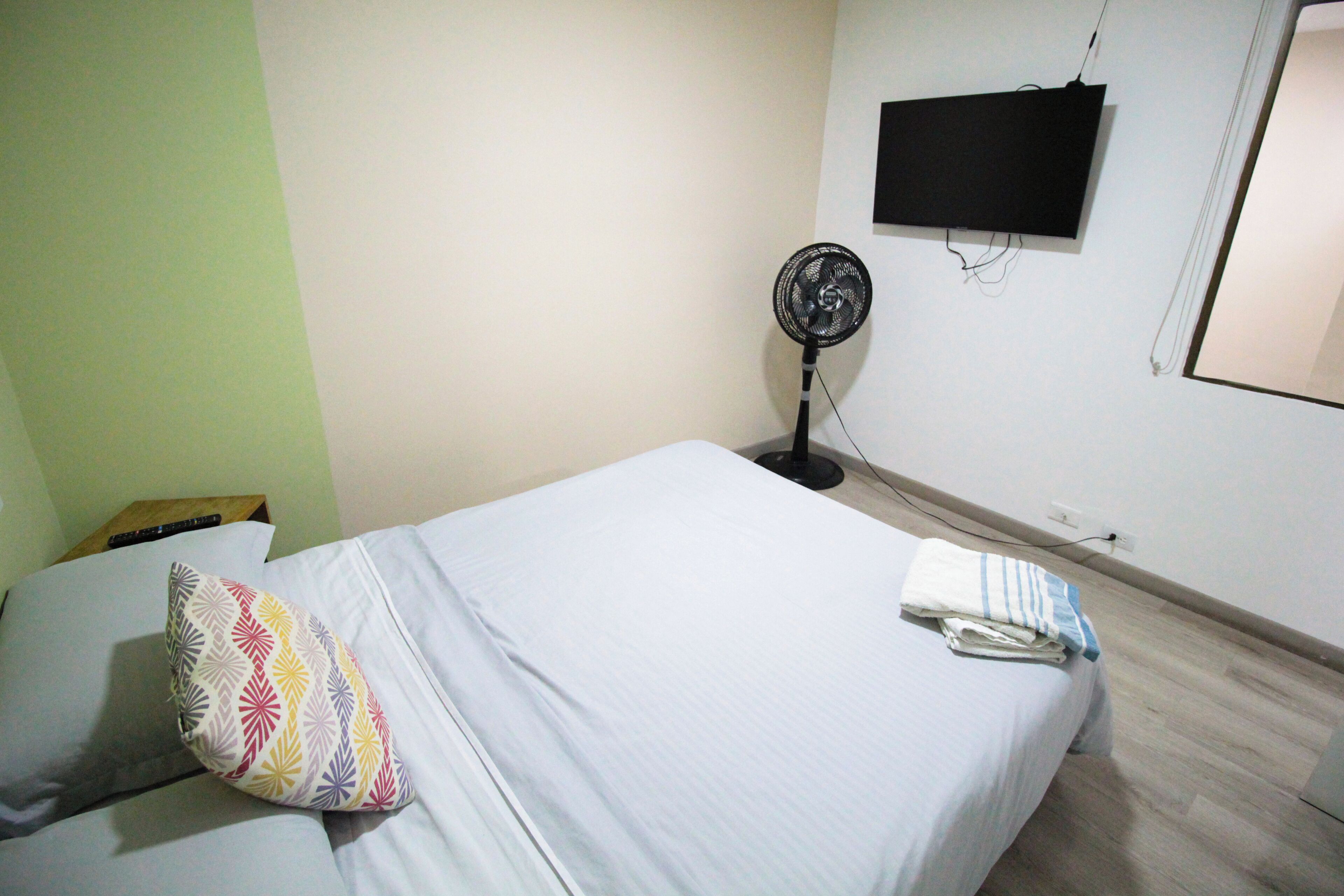 A single bed with a colorful pillow, a white blanket, and a folded towel. A black fan is against the wall, and a TV is mounted above it.