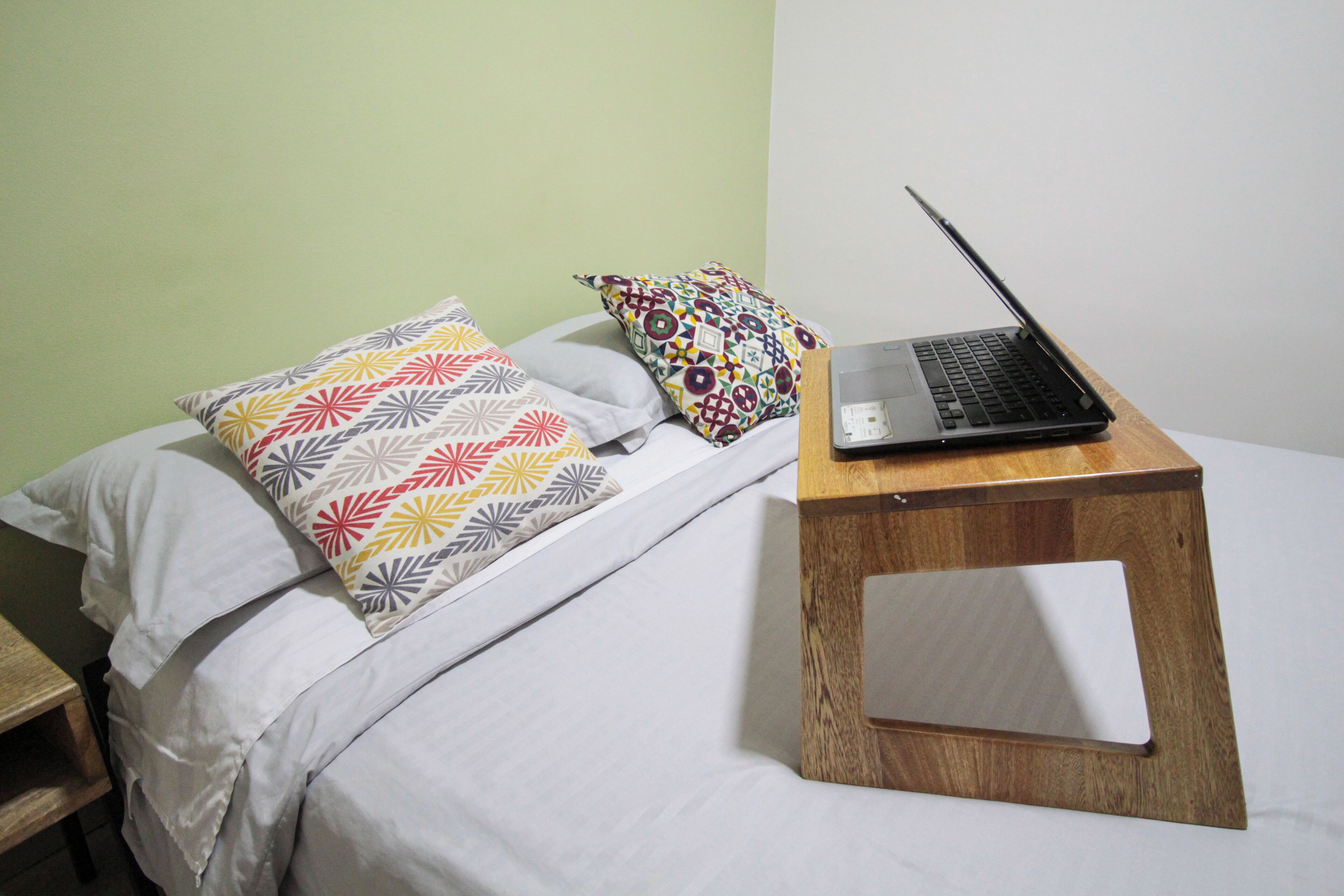 Uma cama com almofadas estampadas, um laptop sobre um suporte de madeira e uma parede lisa.