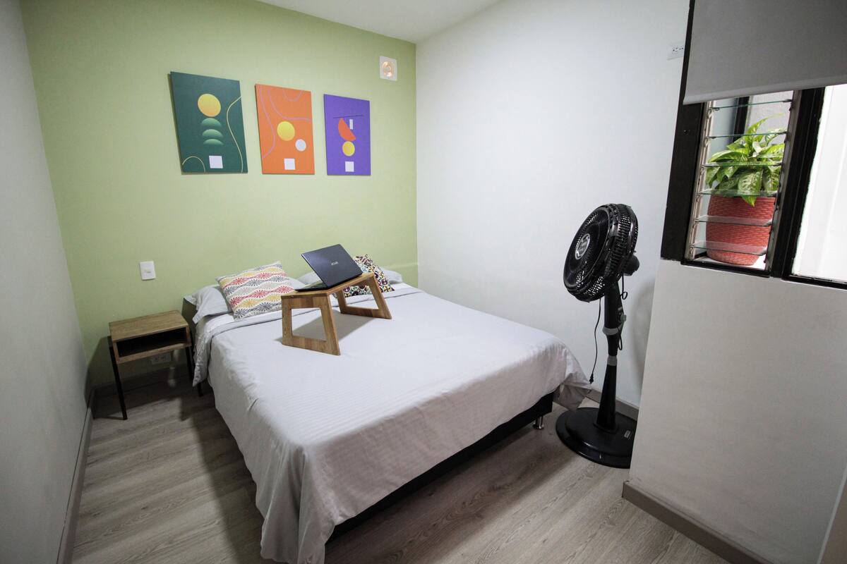 A bedroom with a bed, a laptop on a wooden stand, a standing fan, and three wall art pieces.