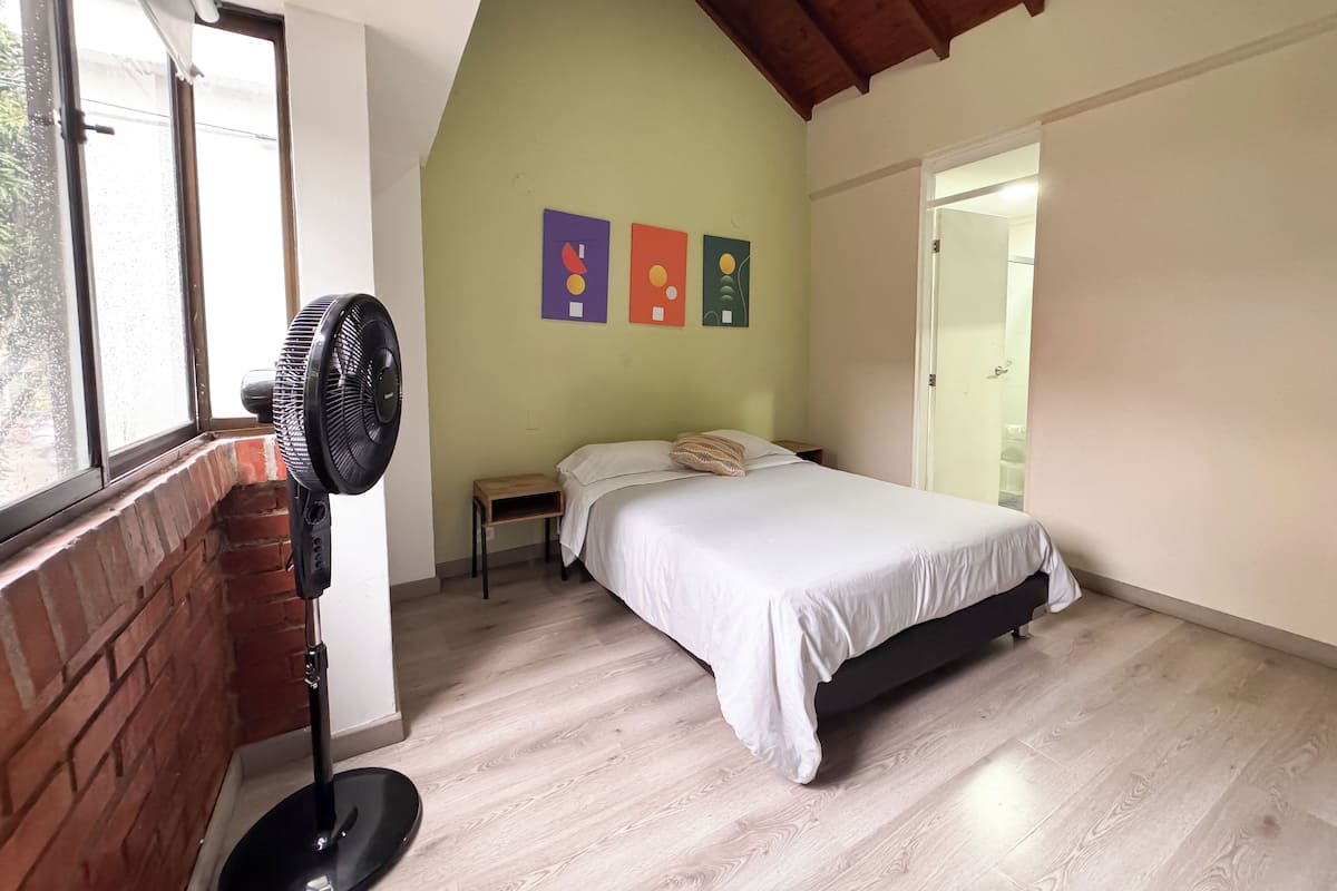A bedroom with a bed, a standing fan, a small table, and a window with a view of greenery.