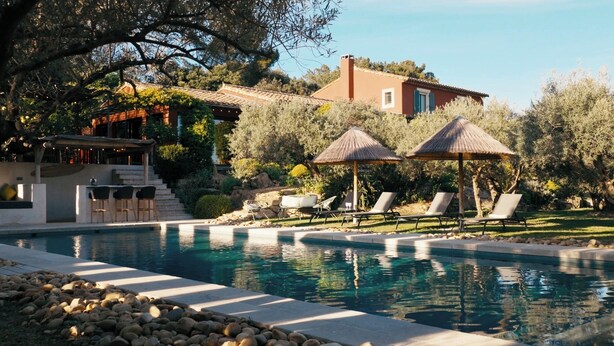 Une piscine avec des chaises longues et des parasols, une maison avec un toit en tuiles et une végétation luxuriante tout autour.