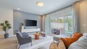 A modern living room with a flat-screen TV mounted on the wall, a grey sofa, a coffee table, and a dining area with a table and chairs. - TR17145BD - 3 bedrooms home close to Disney (Clermont)