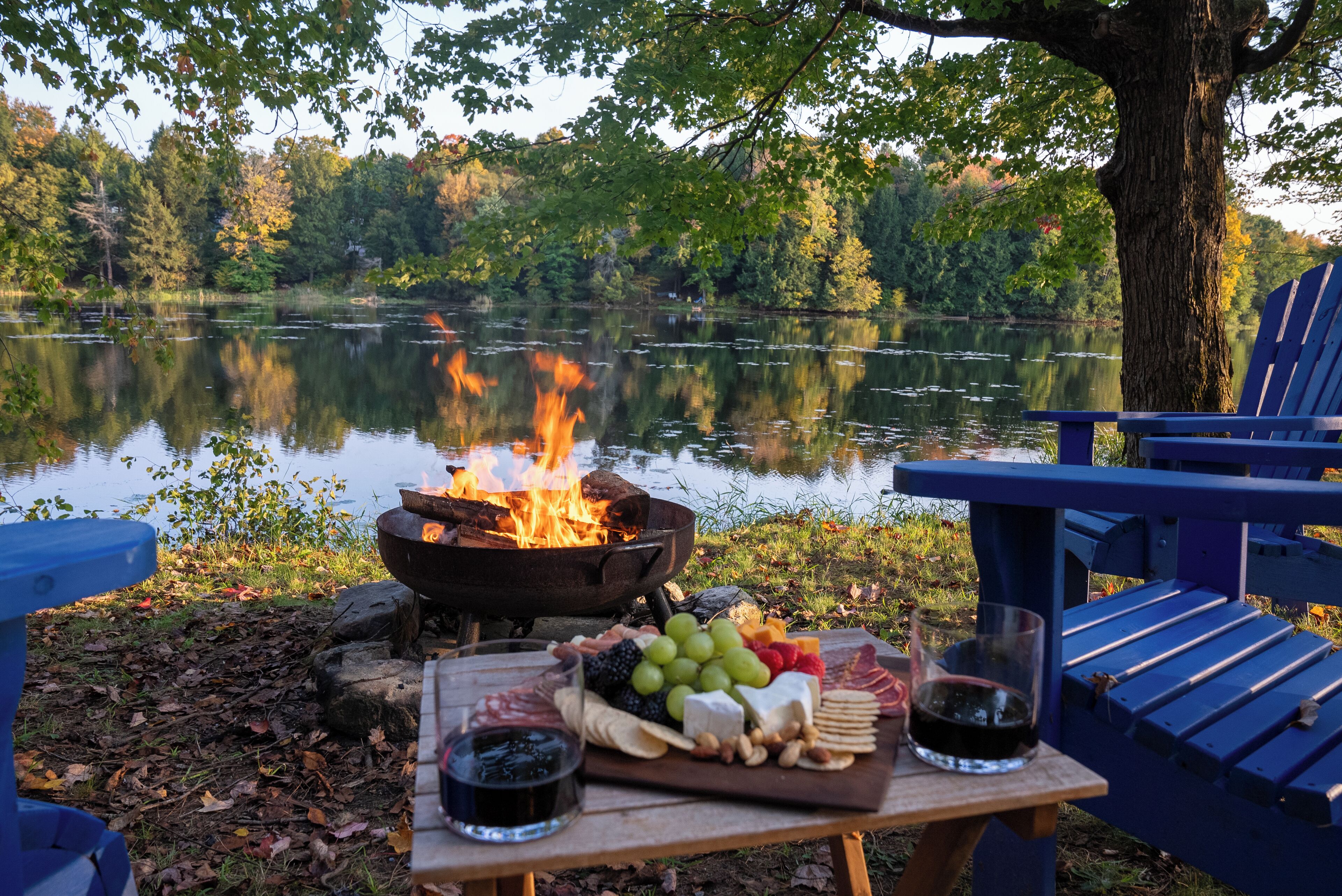 Een picknick aan het meer met een vuurplaats, een tafel met eten en drinken, en een blauwe Adirondack-stoel.