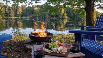 Een picknick aan het meer met een vuurplaats, een tafel met eten en drinken, en een blauwe Adirondack-stoel.