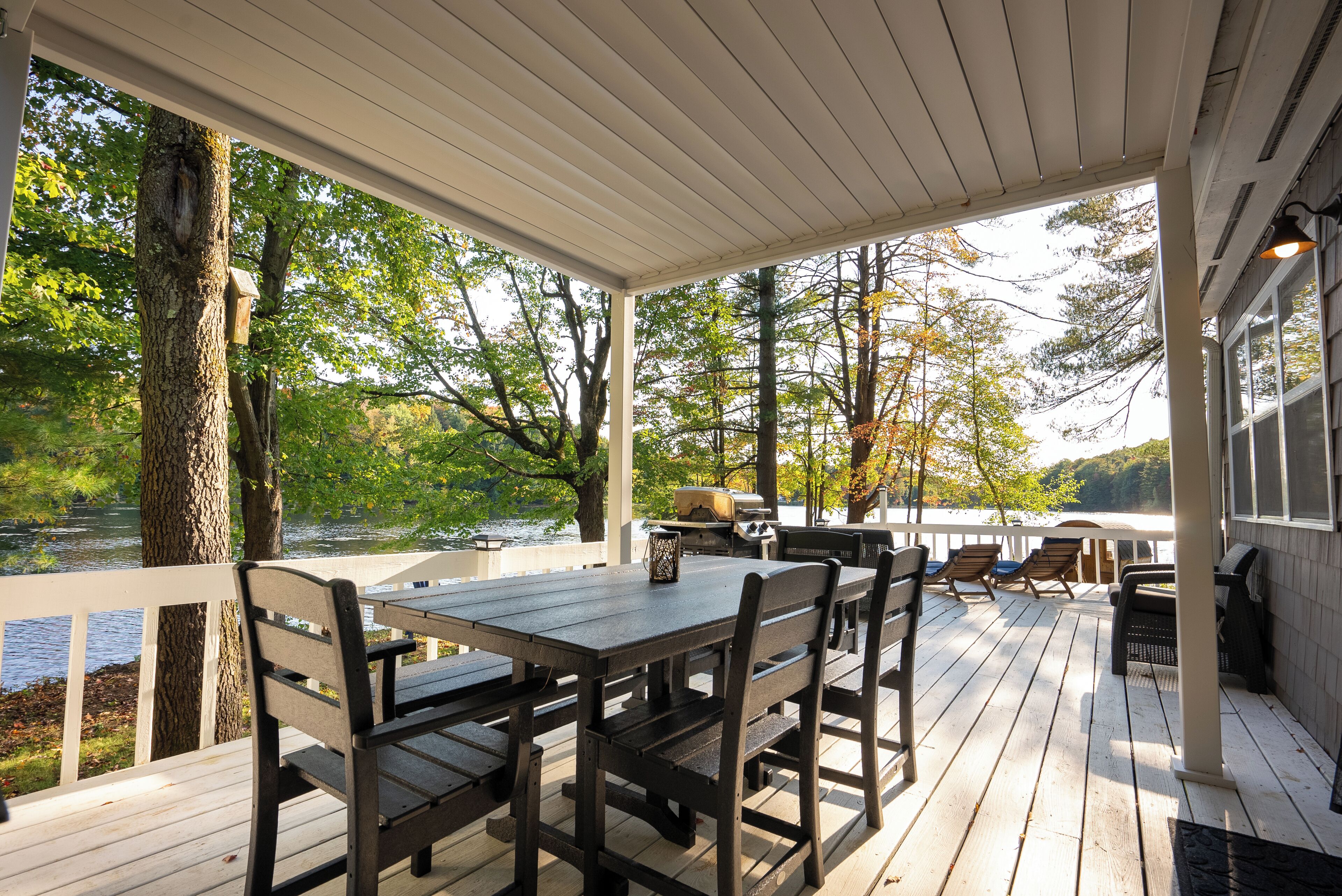 Um deck coberto com mesa e cadeiras, com vista para um lago com árvores e uma churrasqueira.