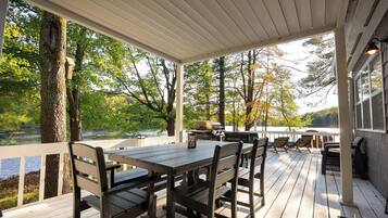 Um deck coberto com mesa e cadeiras, com vista para um lago com árvores e uma churrasqueira.
