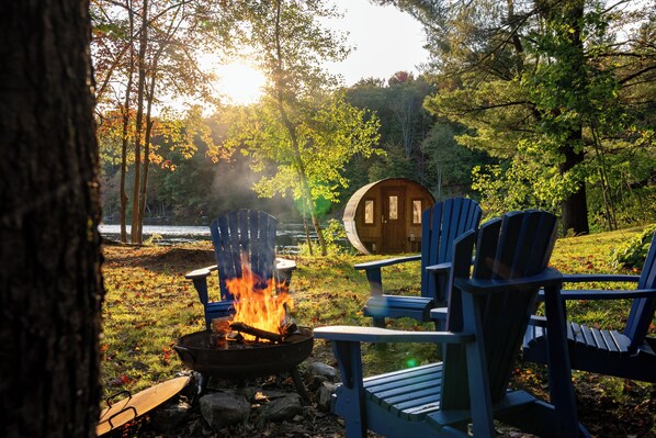 A campfire with chairs around it, a barrel, and trees in the background.