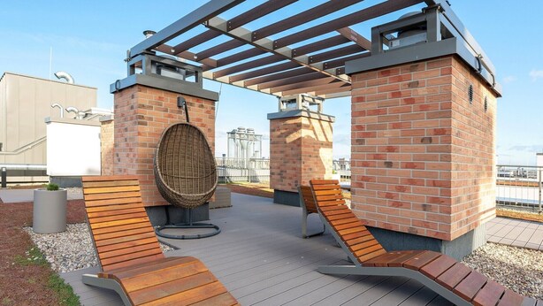 An outdoor seating area with wooden benches and a wicker chair, surrounded by brick pillars and a pergola structure. - Garden Gates by Grand Apartments (Gdansk)