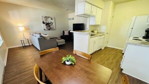 A compact living space with a dining table, a sofa, a kitchen area, and a small side table with a lamp.