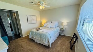 A bedroom with a bed, a ceiling fan, a nightstand with a lamp, and a window with blinds.