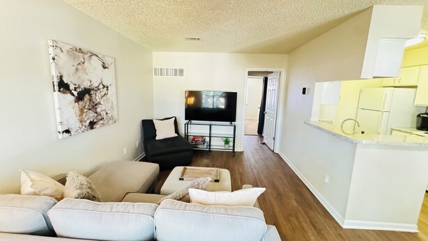 A modern living room with a sofa, a black chair, a TV, and a kitchen area with white cabinets.