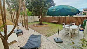 Un patio trasero con cerco de madera, un árbol, un banco, una mesa con sillas y un paraguas verde.