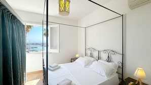 Un letto a baldacchino con lenzuola bianche, un comodino con una lampada e vista sul porto attraverso una porta in vetro.