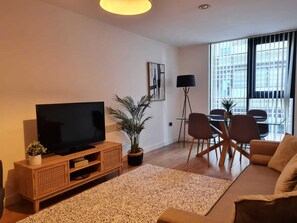Un salon moderne avec un téléviseur à écran plat posé sur une console en bois, une plante en pot et une table à manger en verre avec des chaises.
