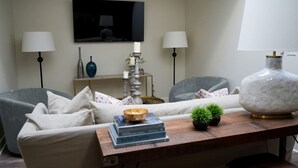 A living room with a sofa, two armchairs, a coffee table with books and a plant, a TV on the wall, and two lamps. - The Nook (Demopolis)