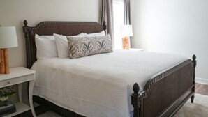 A neatly made bed with a dark wooden headboard, white bedding, and patterned pillows. There are two bedside tables with lamps and a small shelf with books and a vase. - The Nook (Demopolis)