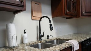 A kitchen with a granite countertop, a stainless steel sink, wooden cabinets, and a wooden cutting board on the wall. - The Nook (Demopolis)