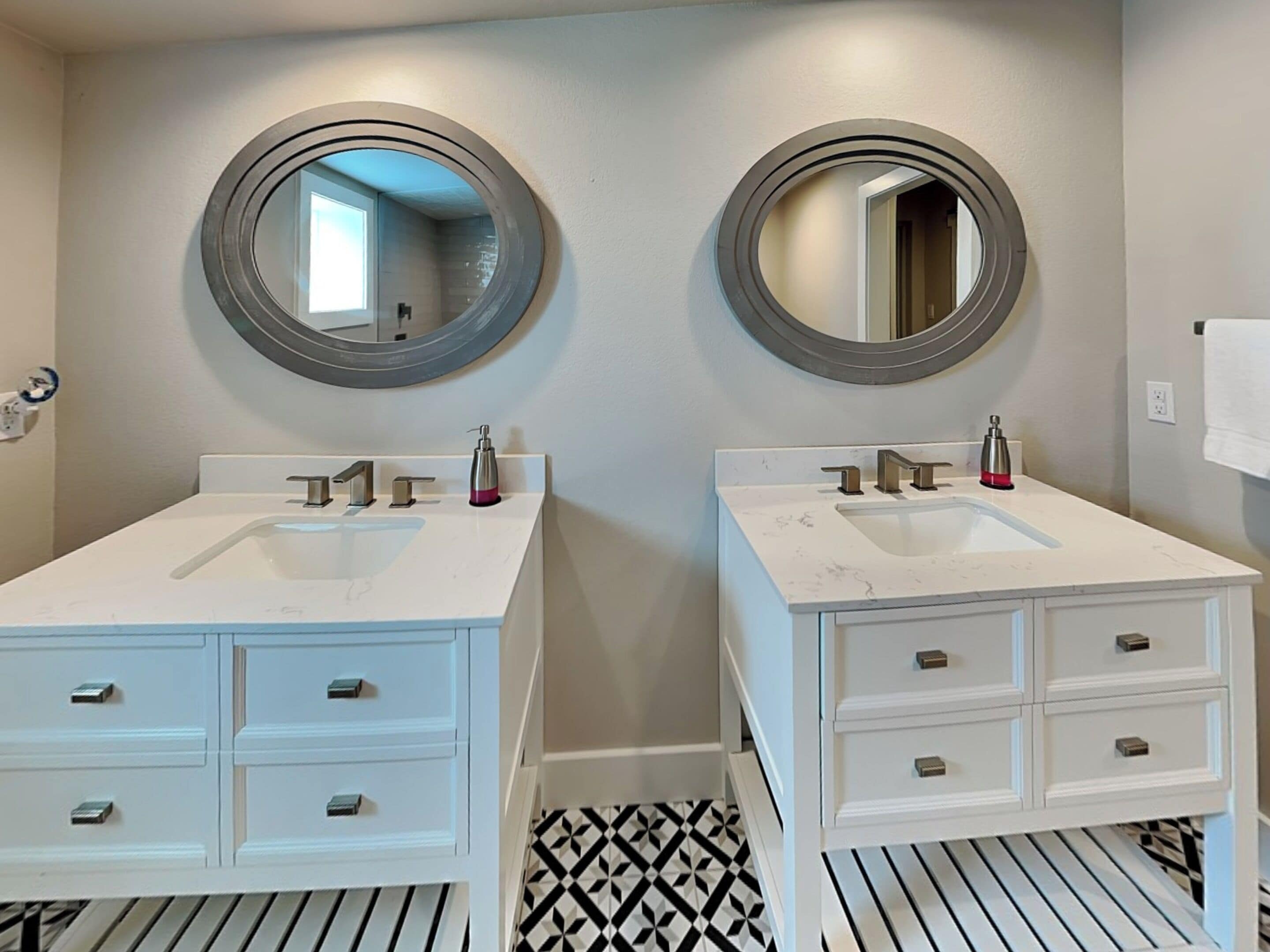Une salle de bain avec deux lavabos, chacun surmonté d’un miroir. Des meubles blancs, dotés de tiroirs situés sous les lavabos, sont également présents. Le sol est recouvert d’un motif géométrique noir et blanc.