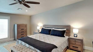 A bedroom with a bed, two bedside tables with lamps, a ceiling fan, a bench, and a window with blinds. - Heron's Roost by Miss Kitty’s (Rockport)