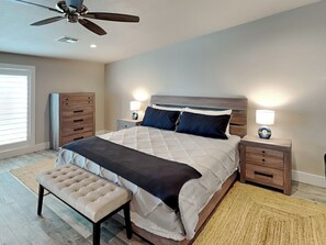 A bedroom with a bed, two bedside tables with lamps, a ceiling fan, a bench, and a window with blinds. - Heron's Roost by Miss Kitty’s (Rockport)