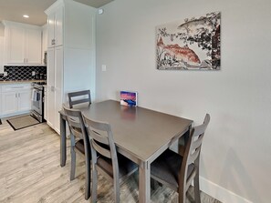 A dining area with a table and chairs, a kitchen in the background, and a painting on the wall.