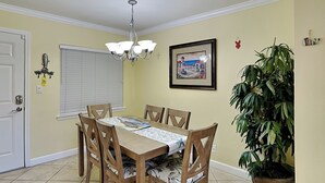 Un comedor con una mesa dispuesta para cuatro personas, rodeada de sillas de madera con cojines estampados. Un candelabro cuelga del techo, y un cuadro enmarcado decora la pared.